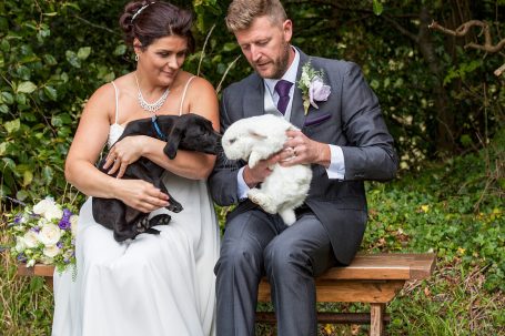 A bride and groom sitting on a bench, holding a black puppy and white rabbit together.