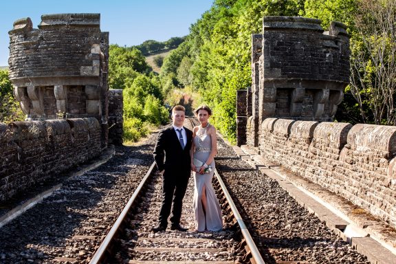 A couple in formal attire standing on railway tracks surrounded by greenery.