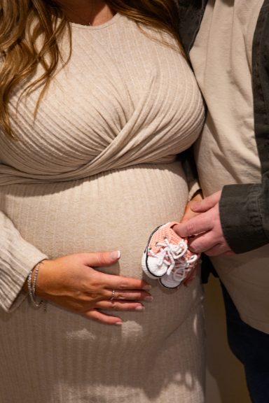 Pregnant person gently holding a pair of baby booties on their belly.