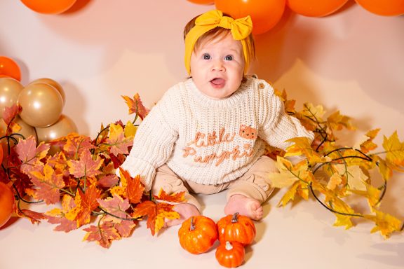 Baby sitting among autumn leaves and pumpkins, wearing a knitted sweater and bow headband.