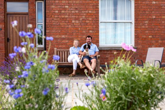 A man and a woman sit on a bench with their dog, surrounded by colourful flowers.