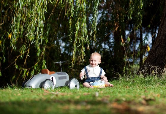 A baby sits on grass near a silver toy car under a tree.