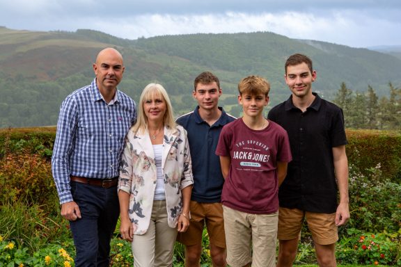 A family of five stands outdoors with mountains and greenery in the background.