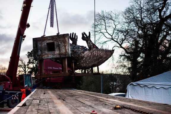 A crane lifts a dilapidated boat onto a flatbed truck against a cloudy sky.
