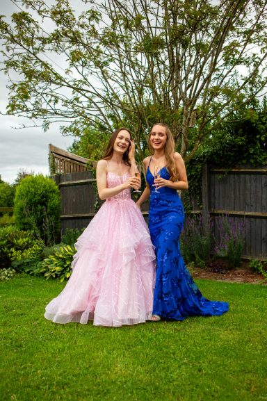 Two women stand in a garden, one in a pink dress and the other in a blue gown.