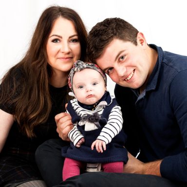 A couple poses with their baby, all smiling against a plain white background.