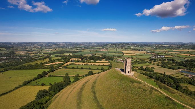 Aerial view of a hill with a stone tower surrounded by green fields and blue sky.