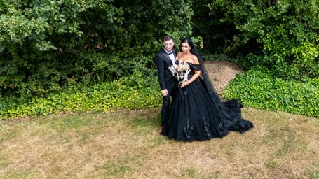 A couple in formal attire stands together, surrounded by greenery. The woman wears a black gown.