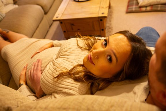 A pregnant woman lies on a sofa, gazing at the camera with a soft smile.