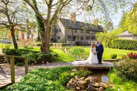 A bride and groom stand on a stone bridge in a lush garden setting.