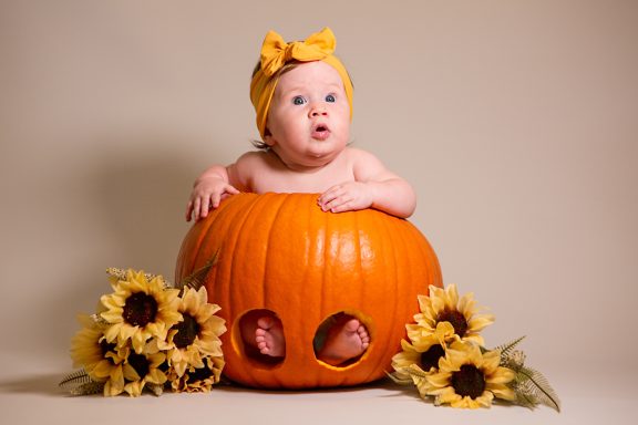 Baby in a pumpkin surrounded by sunflowers, wearing a yellow bow headband.