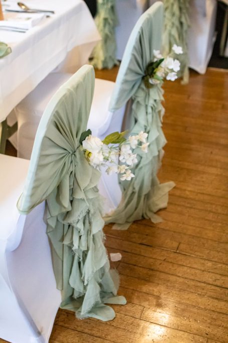 Two chairs draped in light green fabric, decorated with white orchids and flowing fabric.
