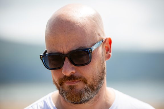 Bald man in sunglasses looking down, with a blurred outdoor background.