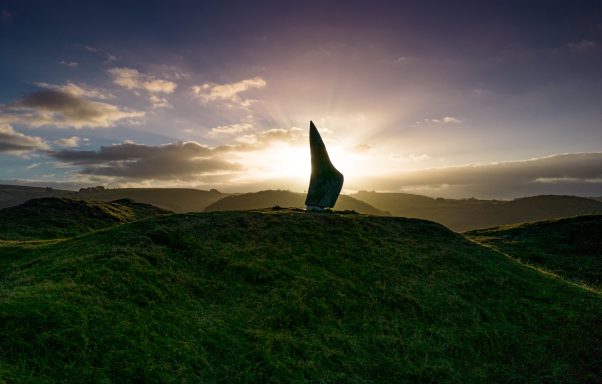 A tall, slender stone monument on a grassy hill at sunset, with clouds and a glowing sky.