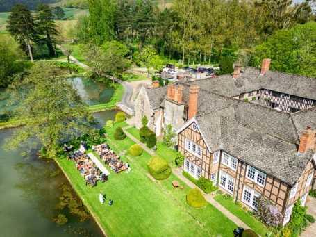 Aerial view of a large house by a pond, surrounded by greenery and outdoor seating.
