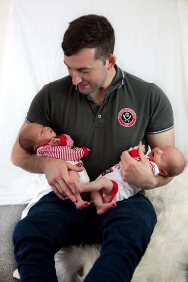 A man sitting, holding two infants in his arms, smiling at them.