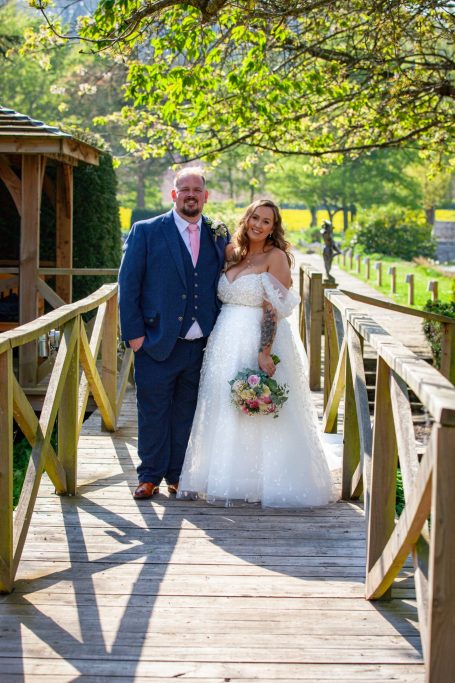 A bride and groom stand on a wooden bridge, smiling, surrounded by greenery.