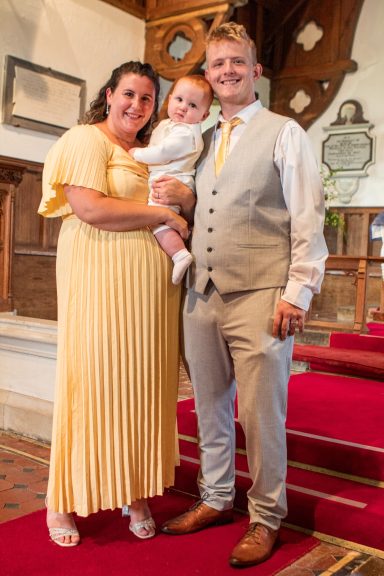A family of three: a couple with a baby, dressed smartly, standing on a red carpet in a church.