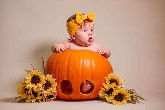 A baby in a pumpkin surrounded by sunflowers, wearing a yellow bow headband.