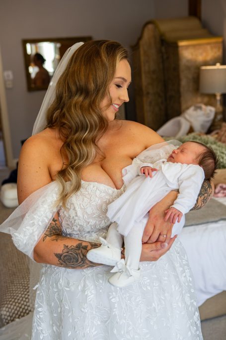Bride in a white gown holding a newborn, smiling warmly in a softly lit room.