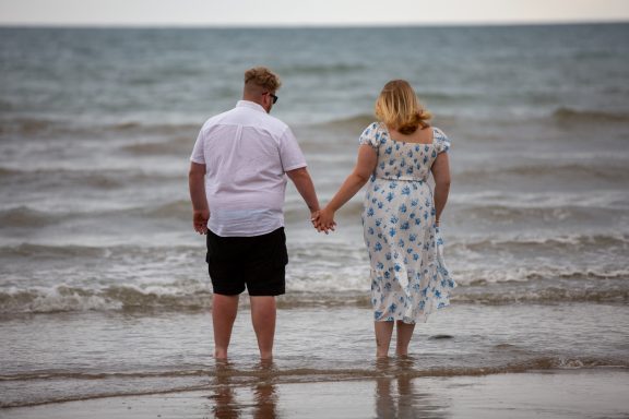 A couple walking hand in hand into the sea, wearing summer outfits.
