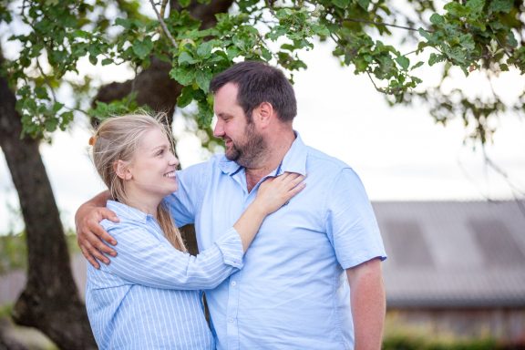 A couple smiling at each other under a tree in a casual outdoor setting.