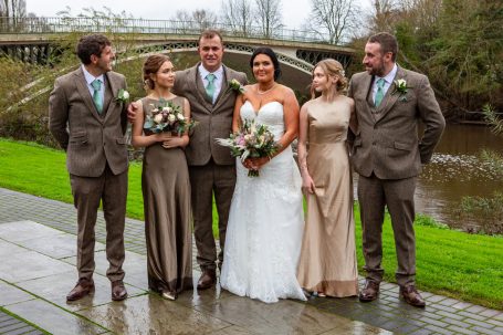 Bride in white dress with bridesmaids and groomsmen pose by a riverbank and bridge.