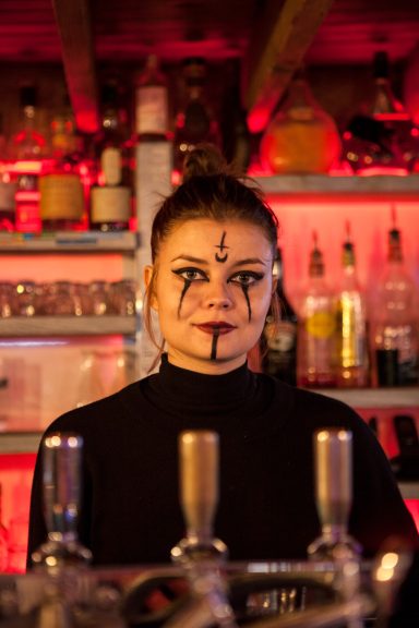 A bartender with face paint stands in front of a well-stocked bar with ambient lighting.