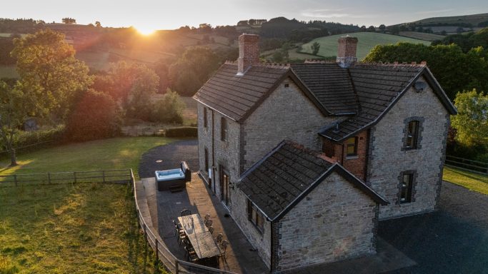 Sunset over a stone house with a garden, surrounded by rolling hills.
