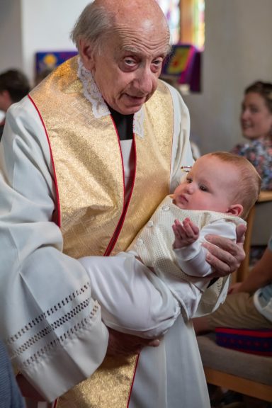 Elderly priest holding a swaddled baby during a ceremony, with people in the background.