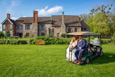 A newlywed couple sitting on a golf cart in front of a stone house and green garden.
