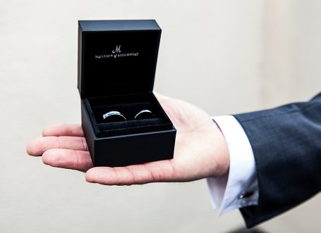 A hand holding a black ring box containing two rings.