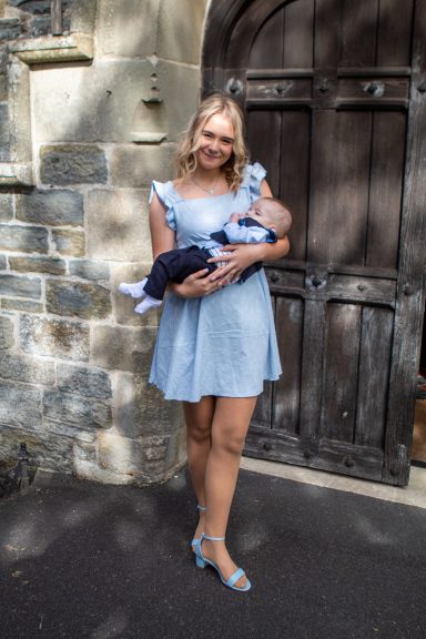 Woman in a blue dress holding an infant, standing in front of a stone wall and wooden door.