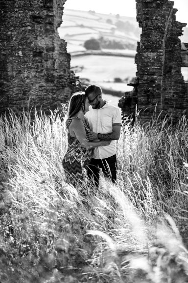 A couple stands close together in long grass, framed by stone ruins.