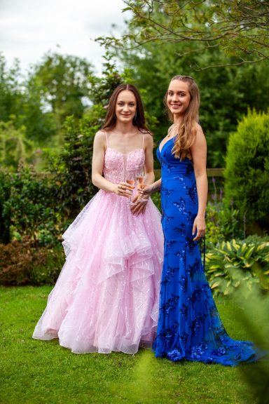 Two young women stand in a garden; one in a pink dress, the other in a blue gown.