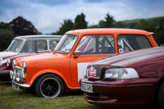 Two classic cars parked in a grassy area, one bright orange and the other dark red.