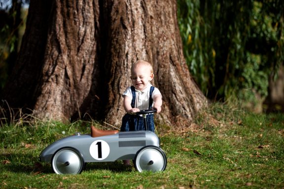 Toddler with a toy car smiling near a large tree trunk.