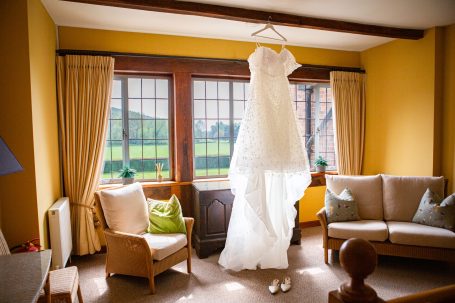 A wedding dress hangs in a well-lit room, with a view of green fields outside.