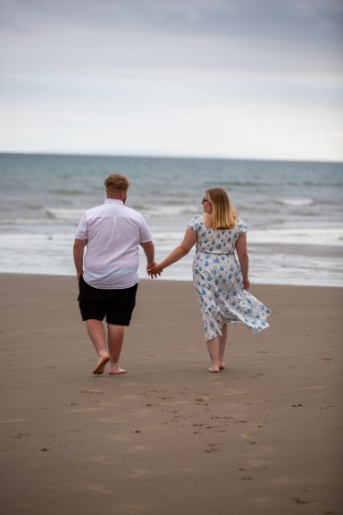 A couple walks hand in hand along a sandy beach with gentle waves.