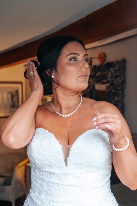 Bride adjusting her hair, wearing a strapless wedding dress and pearl jewelry.