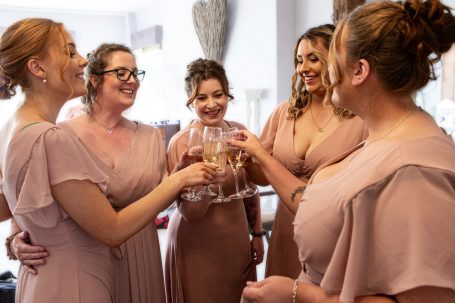 Bridesmaids in blush dresses toasting with glasses in a celebratory setting.