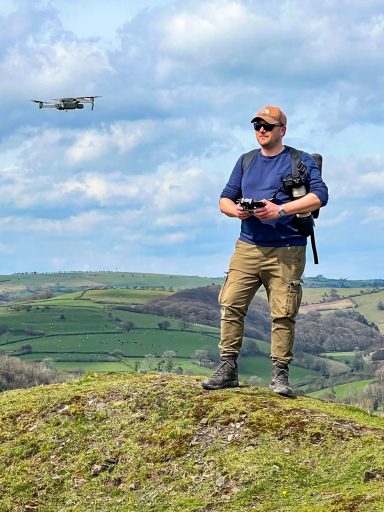 Person in military-style clothing operating a drone on a hillside with a lush landscape.