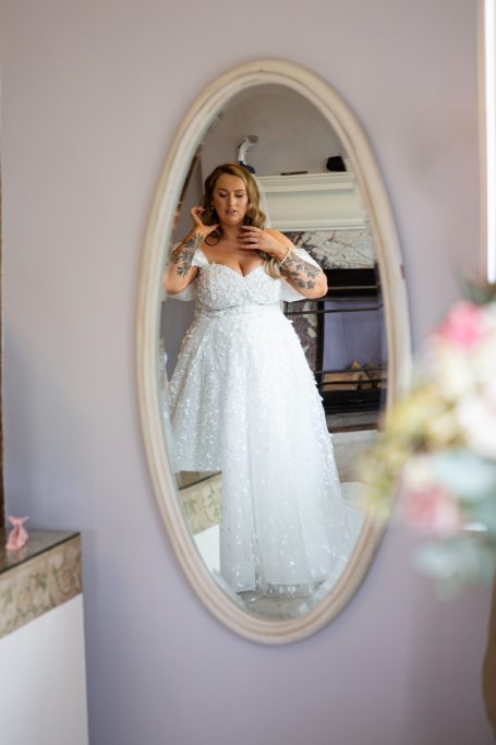 Bride in a white wedding dress adjusting her hair in a mirror with a purple wall backdrop.