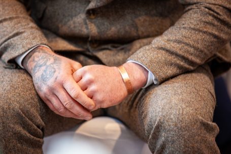 A close-up of a man's hands resting on his lap, wearing a brown suit and a bracelet.