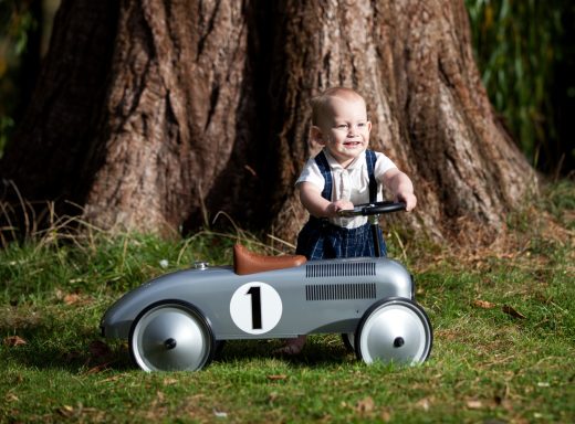 A toddler standing by a tree, holding the steering wheel of a grey toy car.