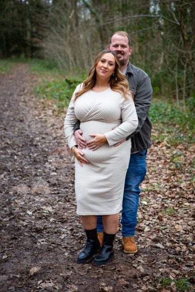A couple poses together outdoors, with the woman cradling her baby bump.