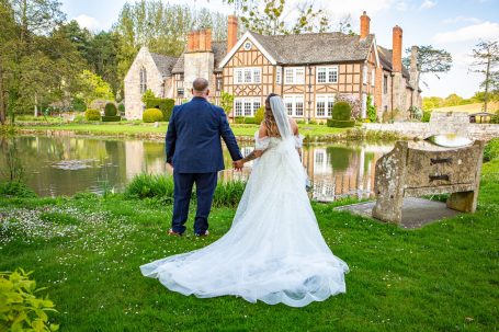 Bride and groom holding hands by a pond in front of a country house.