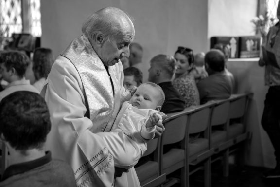 A clergyman holds a swaddled baby during a ceremony, with attendees in the background.