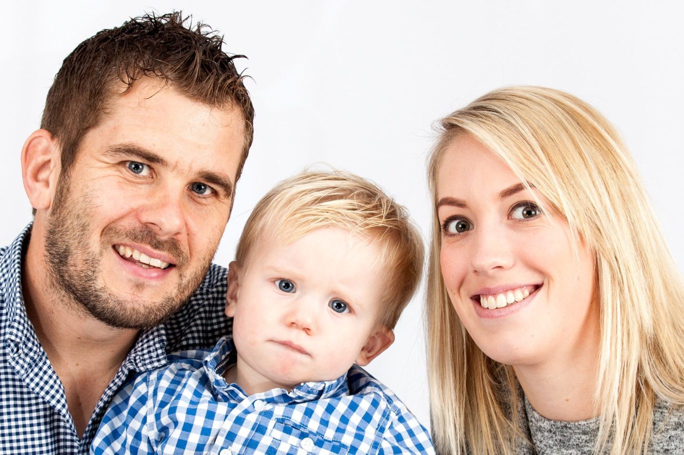 Smiling family portrait with a man, woman, and toddler against a light background.
