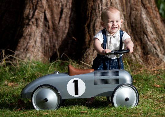 A young child in a denim outfit plays with a vintage toy car outdoors.
